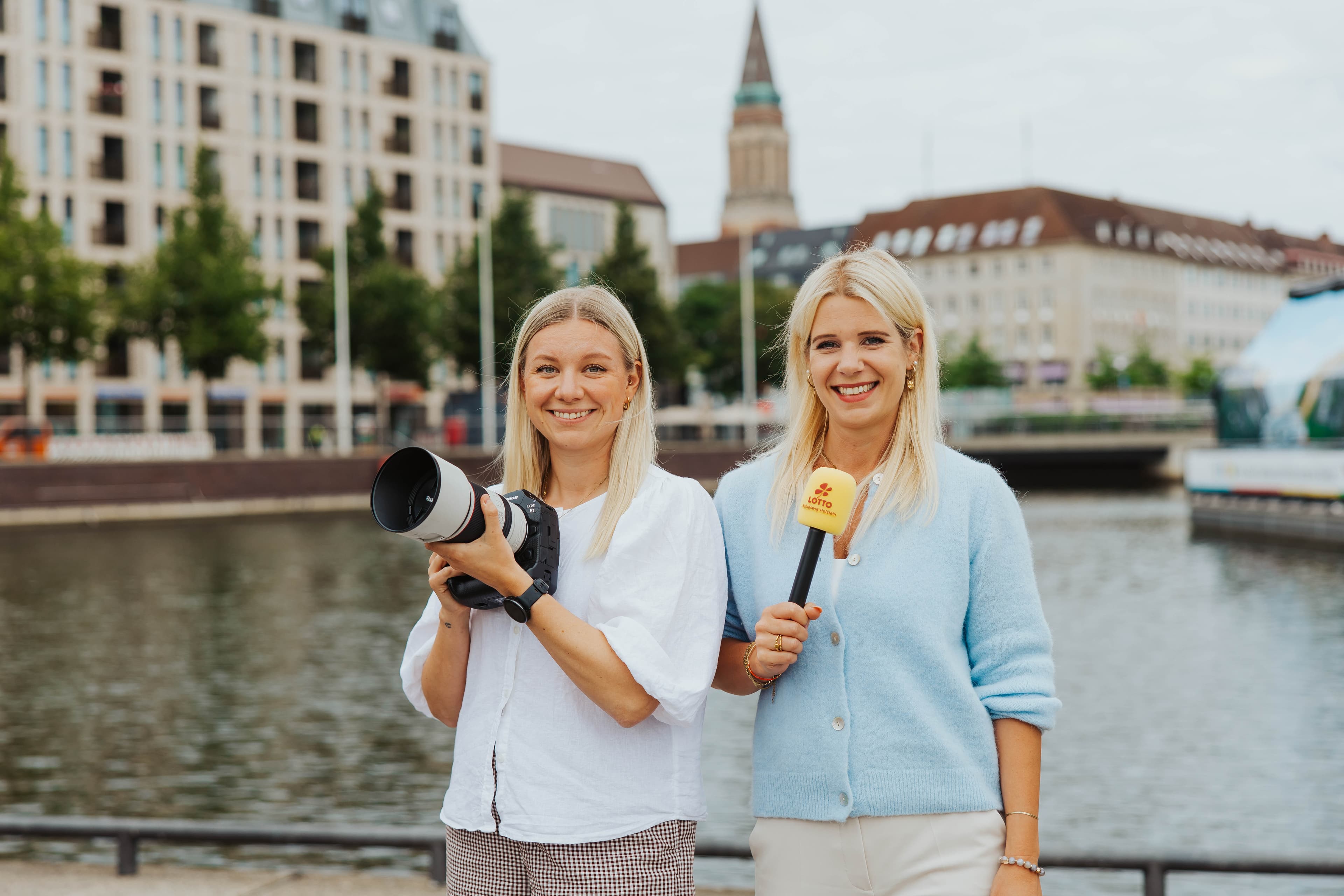 Zwei Frauen lächeln an einer Uferpromenade. Eine hält eine Kamera, die andere ein Mikrofon. Im Hintergrund sind ein Gebäude und ein Kirchturm zu sehen.